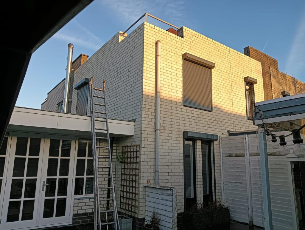 Hoe Herken je een Professionele en Ervaren Dakdekker: Red Flags en Must-Haves 6 Wit bakstenen huis met een ladder aan de zijkant en zonlicht dat op de muur valt, ideaal voor woningbouw en architectuur gerelateerde inhoud.